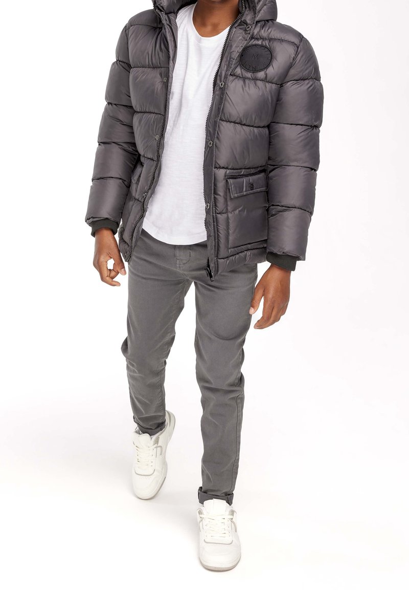 Osoba w szarej kurtce puchowej, białej koszulce, szarych spodniach i białych sneakersach idąca na białym tle.