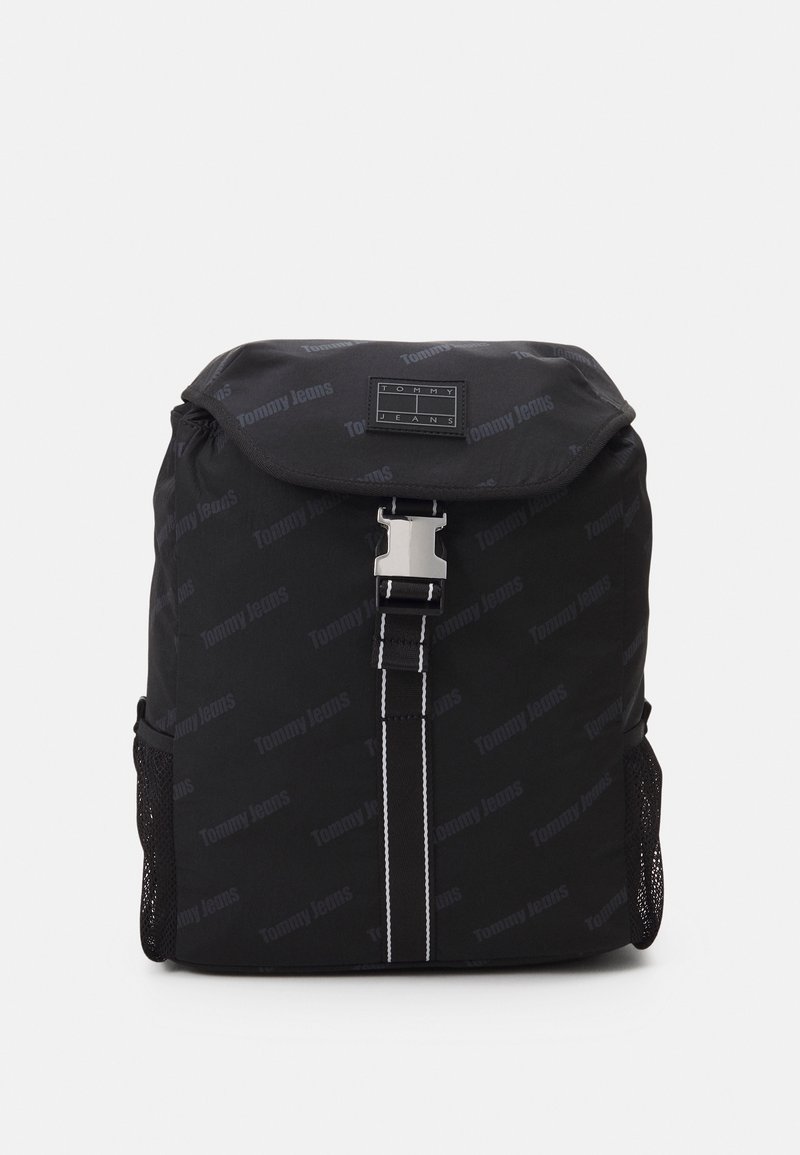 Black Tommy Jeans backpack with flap top, front metal buckle, white stitched vertical strap, and mesh side pockets.