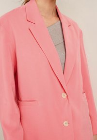 Blazer rosa con un diseño entallado, solapas de muesca, cierre de dos botones y bolsillos laterales. El material parece suave y ligero.