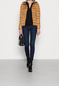 Giubbotto puffer color beige con cappuccio, indossato sopra una blusa nera, abbinato a jeans skinny blu scuro e stivaletti neri; tiene in mano una borsa nera.