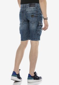 Hombre vestido con pantalones cortos cargo de mezclilla azules de longitud media con parches y cremalleras, zapatillas negras con patrones de estrellas y pulseras de cuentas.