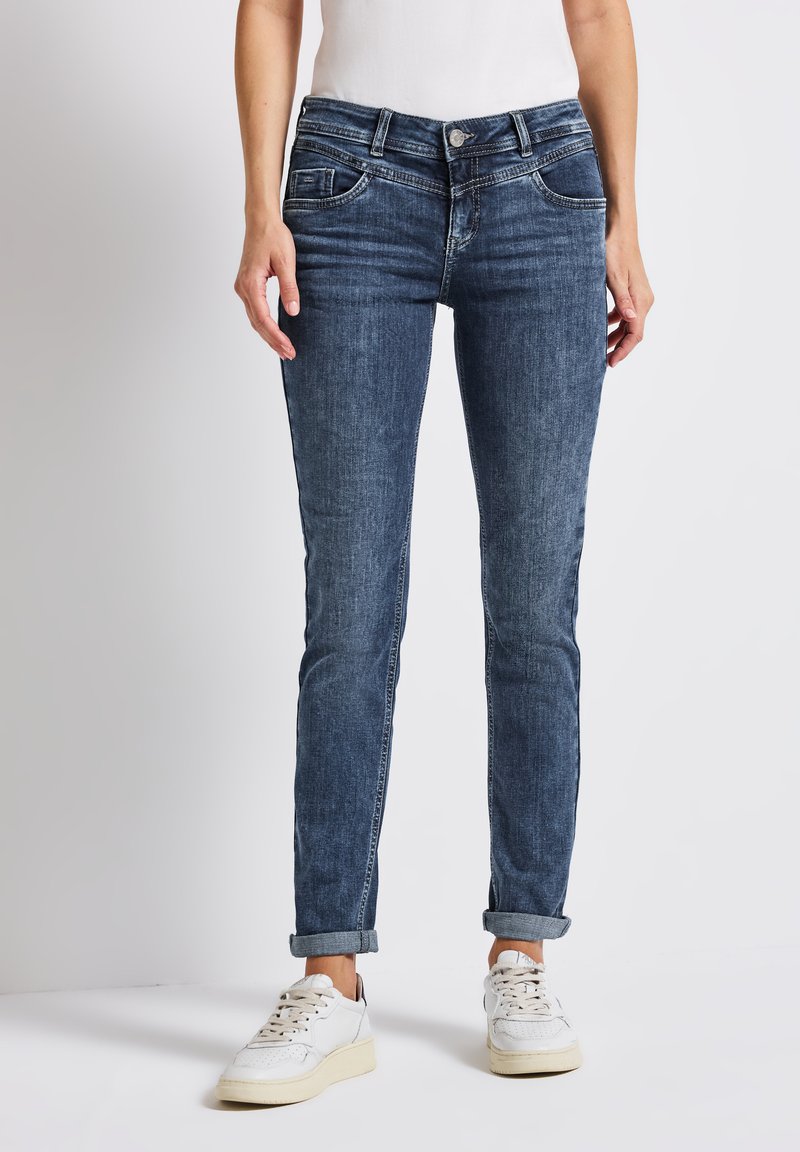 Jeans de mezclilla en azul oscuro con un corte ajustado, diseño de cinco bolsillos y dobladillo vuelto. La tela tiene un ligero desgaste y un detalle de costura visible.