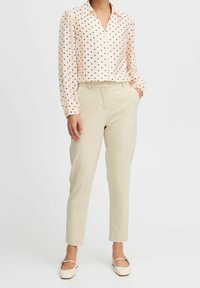Blusa rosa chiaro con pois neri, abbottonata e a maniche lunghe, abbinata a pantaloni beige su misura e scarpe basse di colore chiaro.