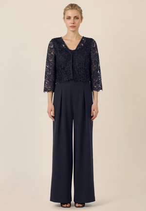 Frau trägt eine marineblaue, aus Spitze gefertigte, kurze Strickjacke mit Dreiviertelärmeln über einem marineblauen Jumpsuit mit weitem Bein und offenen Absatzschuhen.