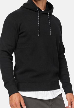 Homme portant un sweat à capuche noir avec cordons blancs, superposé sur un t-shirt blanc, associé à un pantalon gris.