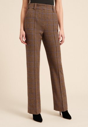 Vrouw draagt hooggetailleerde rechte bruine geruite broek met paarse en zwarte accenten, gecombineerd met zwarte enkellaarsjes met puntige neus.