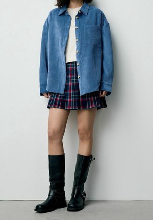 Persona che indossa una giacca camicia blu con bottoni, top bianco, gonna plissettata a quadri e stivali neri al ginocchio, in piedi contro una parete chiara e liscia.