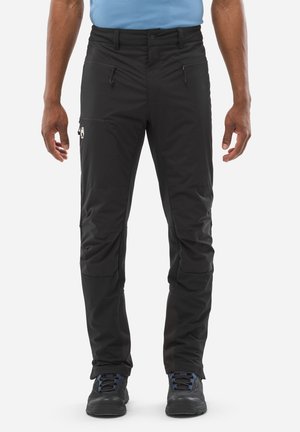 Homme portant un pantalon d'extérieur noir avec des poches zippées et des chaussures de randonnée noires, se tenant contre un fond blanc.