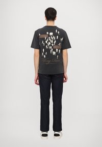 Black t-shirt with a graphic design on the back featuring small human silhouettes and brown accents. Paired with dark denim jeans and sneakers.