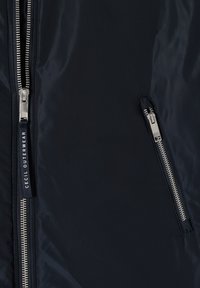Gros plan sur le tissu d'une veste bleu marine montrant deux fermetures éclair argentées, l'une verticale avec un curseur marqué "CECIL OUTERWEAR" et l'autre diagonale pour une poche.