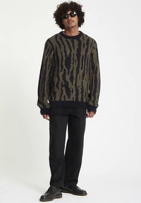 Maglione lavorato a maglia con motivo verde e blu navy, collo rotondo, polsini e orlo a coste; indossato con pantaloni e scarpe nere.