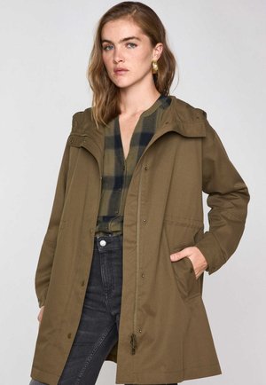 Giacca con cappuccio verde oliva, con zip frontale, bottoni a pressione e tasche laterali. Indossata sopra una camicia a quadri e abbinata a jeans scuri.