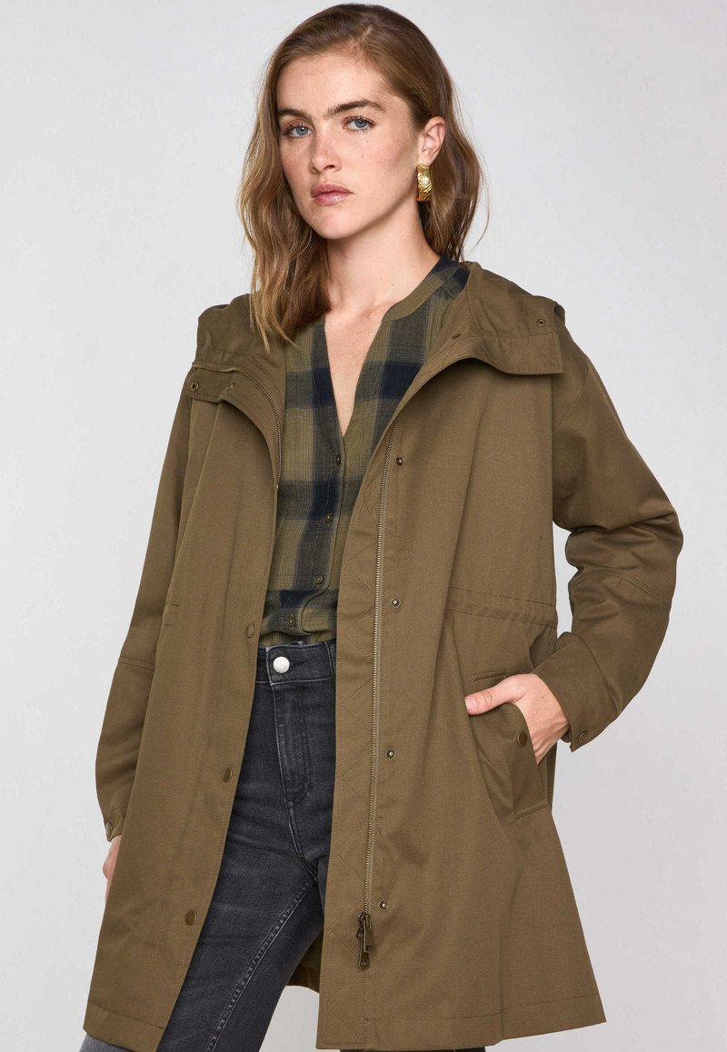 Olivgrüne Kapuzenjacke mit einem Frontreißverschluss, Druckknöpfen und Seitentaschen. Über einem karierten Hemd getragen und kombiniert mit dunkelblauer Jeans.