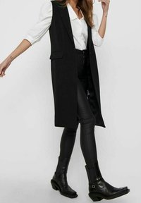 Długi czarny kamizelka z szerokimi klapami, zestawiona z białą koszulą i czarnymi skórzanymi spodniami. Stylizacja uzupełniona czarnymi botkami za kostkę.