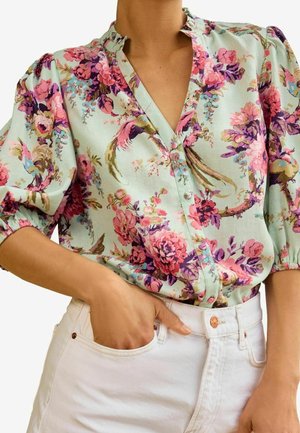 Vrouw draagt een lichtblauw bloesje met bloemenprint en pofmouwen, en een witte hoge broek, hand in de zak, tegen een effen achtergrond.