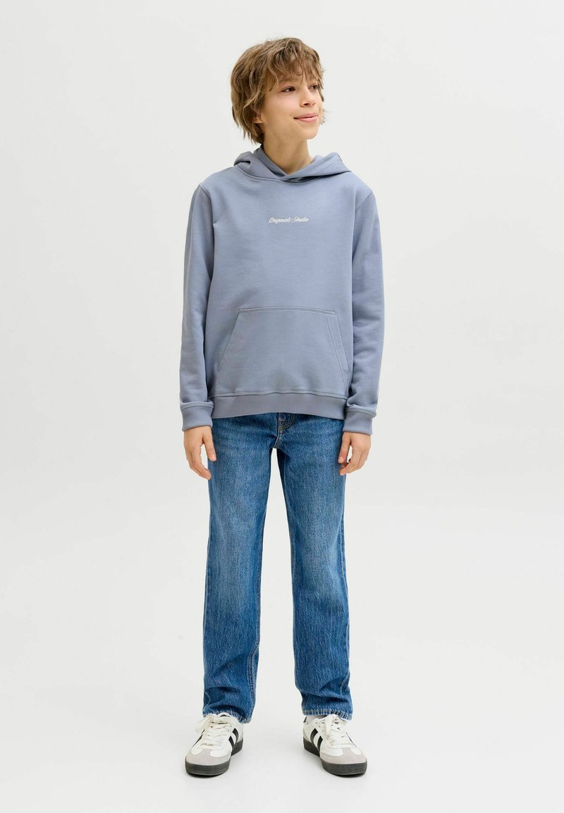 Junge steht, trägt einen hellblauen Kapuzenpullover, blaue Jeans und weiße Turnschuhe, schaut leicht zur Seite vor einem neutralen Hintergrund.