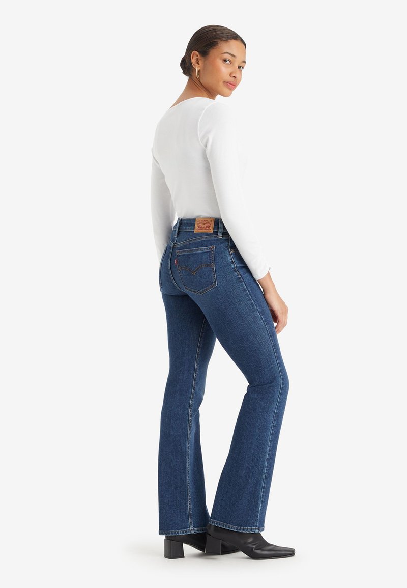 Donna con i capelli raccolti in un bun, che indossa orecchini a cerchio dorati, una maglietta bianca a maniche lunghe, jeans svasati blu e stivali neri con tacco, in posa mentre guarda oltre la spalla.