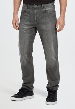 Modèle portant un jean gris foncé à coupe ample, un haut noir et des baskets noires sur un fond clair uni.