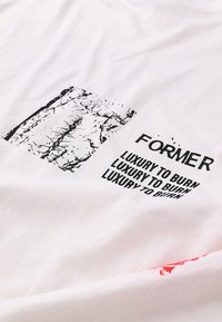 Λευκό βαμβακερό μπλουζάκι με εκτυπωμένο μαύρο κείμενο και γραφικό σχέδιο. Το κείμενο γράφει «FORMER LUXURY TO BURN» με υφή σχεδίου.