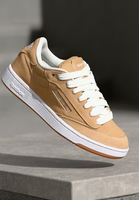 Sneaker in camoscio beige con lacci bianchi e suola in gomma. Presenta un patch con logo e una punta forata. Design semplice con cuciture a contrasto.