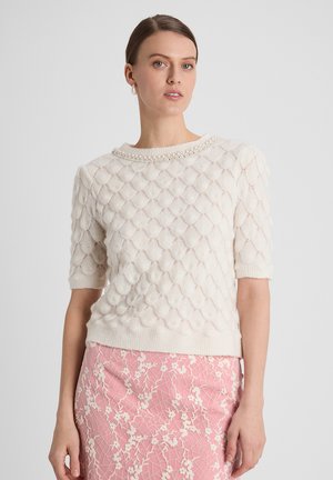 Vrouw draagt een witte, gestructureerde gebreide top met parel halslijn en een roze rok met bloemenpatroon, staand tegen een effen achtergrond.