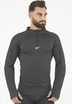 Mann mit Bart, der ein eng anliegendes, langärmliges dunkelgraues Sportoberteil mit Frontreißverschluss und Logo trägt, Hände in der Nähe der Taille, schaut nach rechts.