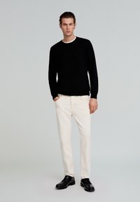 Jersey negro de punto con cuello redondo, pantalones color crema y zapatos de cuero negros. El modelo está de pie recto frente a un fondo neutro.