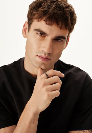 Ung mand med kort brunt hår iført en sort t-shirt og en sort ring, hvilende hagen på hånden, kigger på kameraet.