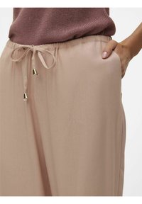 Pantalons roses clairs avec une taille à cordon de serrage, arborant des accents dorés. Texture lisse et coupe décontractée, avec des poches latérales visibles.