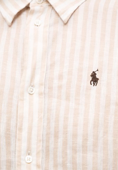 Chemise à rayures verticales beige et blanches, boutonnée, avec un petit logo brodé représentant un joueur de polo en marron foncé sur la poitrine.