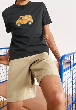 Musta T-paita, jossa keltainen toimituspakettiauton grafiikka; yhdistetty khakinvärisiin shortseihin, asetettuna puiselle pinnalle, jossa sinisiä metallikoristeita.