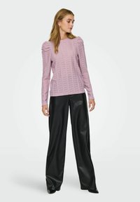 Kvinne iført en rosa blonder langermet topp med puffede skuldre og svarte vide skinnbukser som står mot en ensfarget bakgrunn.