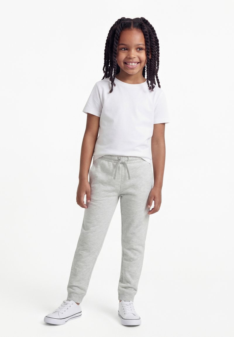 Ragazza sorridente con lunghe trecce che indossa una maglietta bianca a maniche corte, pantaloni della tuta grigio chiaro e scarpe da ginnastica bianche, in piedi su uno sfondo bianco.