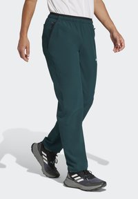 Mørk teal sportsbukser med elastisk talje, tapered design, sidelommer og sorte detaljer; bæres med sorte træningssko.
