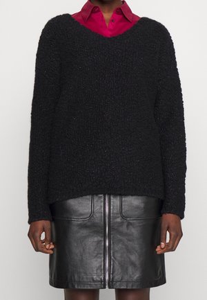 Osoba ubrana w czarny sweter o fakturze na czerwonej koszuli z kołnierzykiem, w połączeniu z czarną skórzaną spódnicą z przednim zamkiem błyskawicznym.