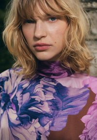 Mladý člověk s vlnitými blond vlasy, oblečený v purpurovém topu s květinovým vzorem a vysokým límcem, dívající se přímo s neutrálním výrazem.