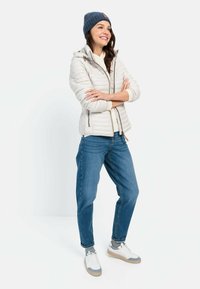 Lettet, hættejakke i cremefarvet puffer-materiale med lynlås foran. Båret med blå denim jeans og hvide sneakers med grå detaljer. Strikket blå hue.