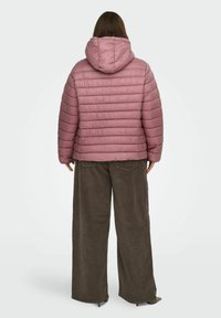 Kvinne iført en lilla quiltet hettejakke og vide mørkebrune kordfløyelsbukser, stående med ryggen til foran en ensfarget bakgrunn.