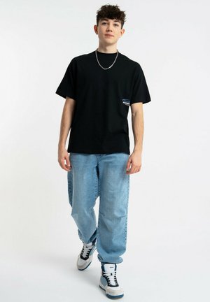 Młody mężczyzna ubrany w czarny t-shirt, jasnoniebieskie luźne dżinsy, białe i niebieskie sneakersy oraz srebrny łańcuszek, idzie do przodu.