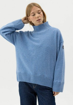 Sieviete, tērpusies gaiši zilā pārlielā augstu apkaklītē džemperī un tumšās džinsās, paceļot vienu roku un novelkot to aiz galvas.