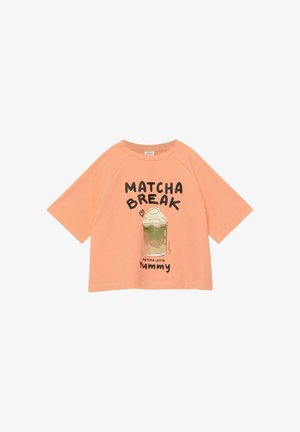 Pfirsichfarbenes Kurzarmhemd mit Aufdruck eines Matcha-Latte, garniert mit Schlagsahne, und dem Text "MATCHA BREAK" sowie "Yummy" auf der Vorderseite.