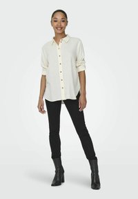 Helles kremfarbiges Hemd mit aufgerollten Ärmeln, kombiniert mit schwarzen engen Jeans und schwarzen, knöchelhohen Stiefeletten mit Absatz. Einfaches Design, ohne Muster.