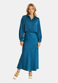 Blusa de manga larga en color verde azulado y falda a juego con adornos dispersos similares a perlas. Tejido suave, cintura entallada, falda fluida. Zapatos blancos.