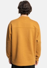 Sudadera amarilla mostaza con cuello redondo, mangas largas y un corte relajado. Presenta una textura suave y una costura horizontal distintiva en la parte posterior.