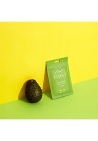 Grön avokado bredvid en grön förpackning för "Avo Cado Nourishing Scalp Pack med Banan" på en ljusgul bakgrund.