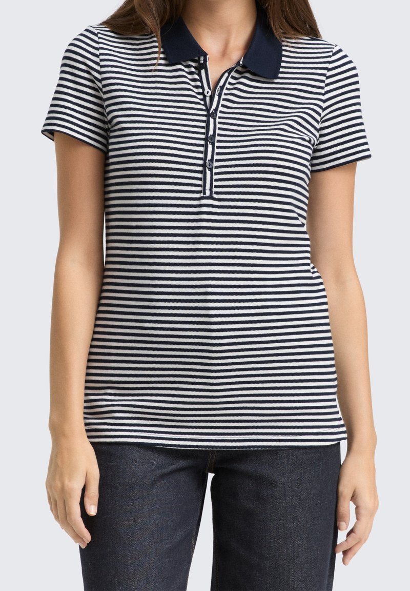 Femme portant un polo à manches courtes rayé horizontalement bleu marine et blanc avec patte de boutonnage et un jean bleu foncé, debout devant un fond uni.