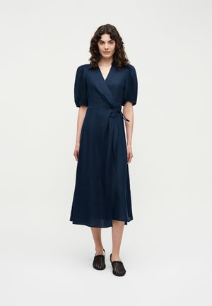 Kobieta stojąca, ubrana w granatową midi sukienkę kopertową z bufkami i czarne plecione wsuwane buty, na jednolitym jasnym tle.