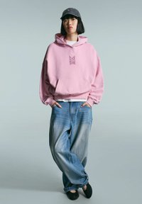 Person stands with hands in pockets wearing a pink hoodie, loose blue jeans, black shoes, and a dark cap against a plain background.
