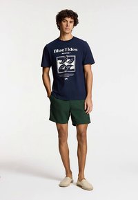 Laevastiku graafiline t-särk tekstiga "BlueTides Resort", rohelised lühikesed püksid ja beežid libisemisjalanõud. Vabaaja sobivus ja lihtne disain.