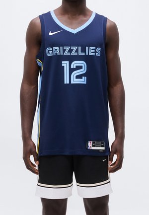 Homme portant un maillot de basketball bleu marine des Memphis Grizzlies avec le numéro 12 et un short noir avec un liseré blanc, debout devant un fond uni.
